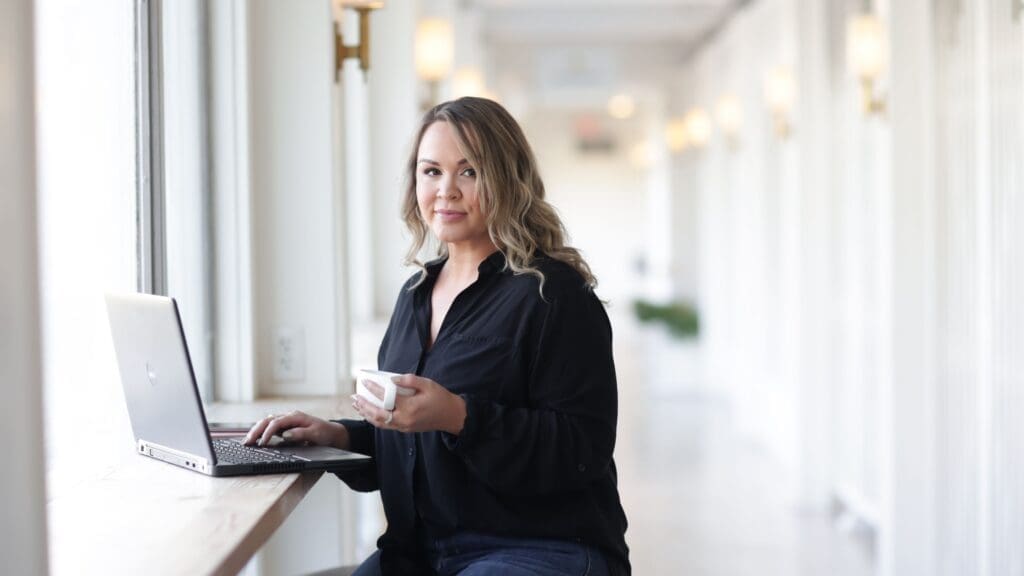 photo of woman sitting at laptop, holding a cup of tea, and smirking